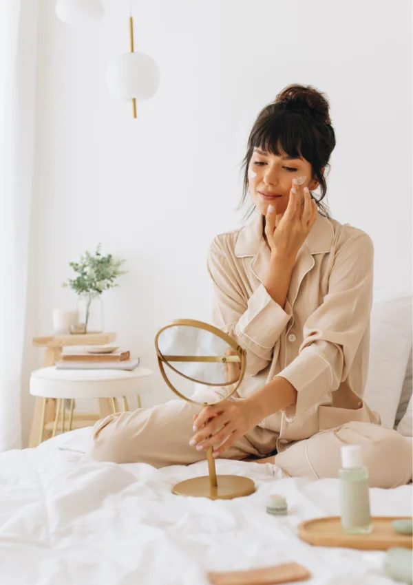 Come combinare i principi attivi. Donna che applica la crema sul viso seduta sul letto come parte della sua routine di cura della pelle.