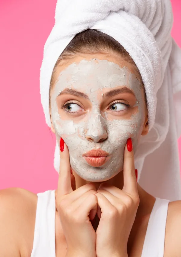 Donna con asciugamano sui capelli e maschera viso all'argilla su sfondo rosa, tocca il viso con espressione giocosa